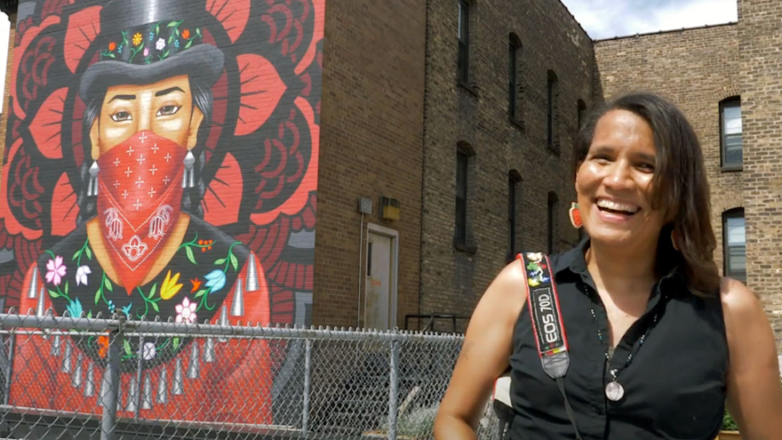 headshot photo of photographer Ivy Vainio standing by AICHO's building with mural of Native American female wearing a red mask in background