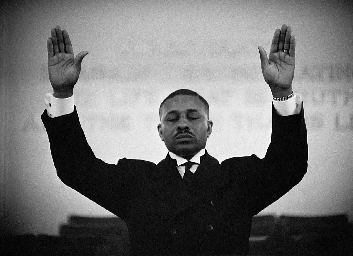 black preacher in white shirt and dark suit with arms up in the air and eyes shut in prayer
